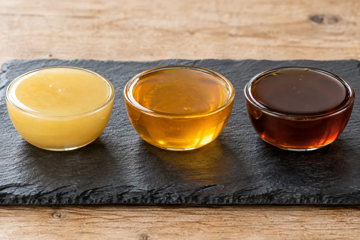 Trois coupelles en verre remplies de miels de couleurs différentes, du jaune pâle crémeux au brun foncé, alignées sur un plateau en ardoise